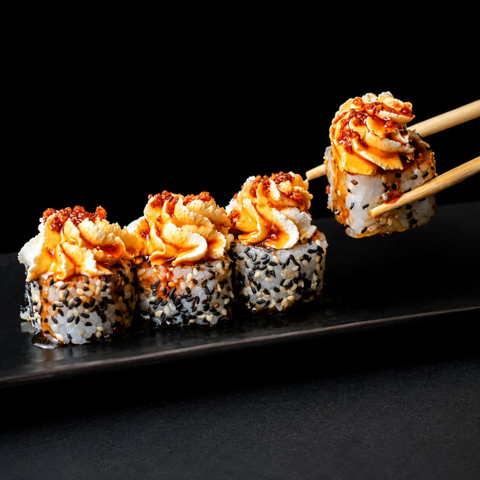 Chopsticks lifting a sesame seed sushi roll topped with creamy sauce and red roe from a black plate.